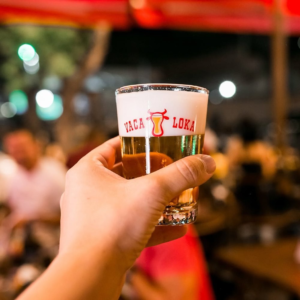 Mão erguendo um copo de chopp com o logo do Vaca Loka, com o bar ao fundo.